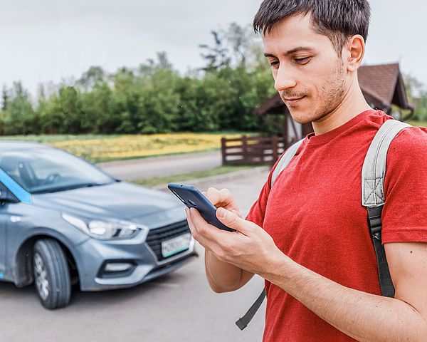 Ein junger Mann steht auf einem Parkplatz neben einem Auto und tippt etwas in sein Smartphone.