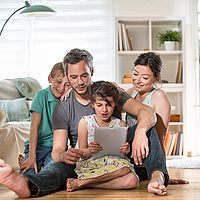 Eine glückliche Familie sitzt am Wohnzimmerboden und blickt voller Spannung auf ein Tablet.