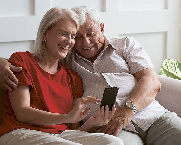 Älteres Paar sitzt auf der Couch und blickt fröhlich auf Smartphone.