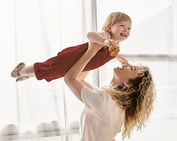 Frau hebt Kind wie ein Flugzeug in die Höhe. Beide lachen.