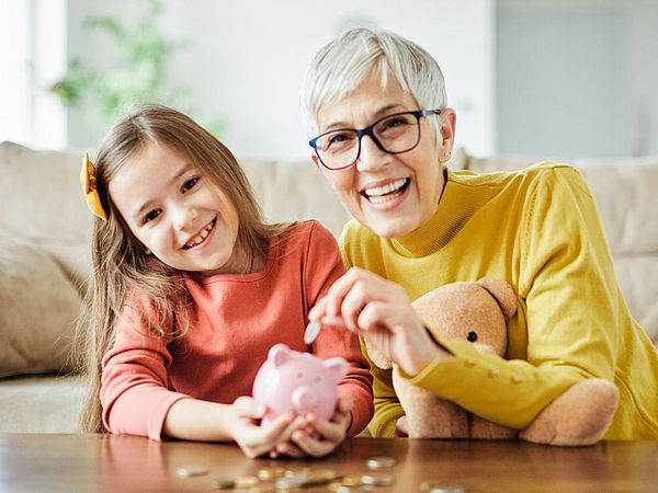 Sehr moderne Oma mit Brille und Kurzhaarschnitt wirft Geld in das Sparschwein ihrer Enkeltochter.
