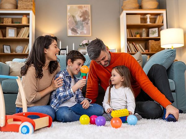 Mutter, Vater und zwei Kinder sitzen zusammen am Boden umringt von Spielsachen.