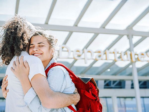 Ein Mann mit langen, gelockten, dunklen Haaren und eine Frau mit dunklen Haaren und rotem Rucksack umarmen sich