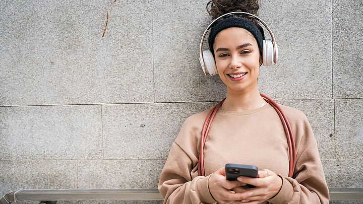 Eine junge Frau mit schwarzen, lockigen Haaren lacht in die Kamera. Sie trägt weiße Over-Ear-Kopfhörer und hält ein Smartphone in beiden Händen.