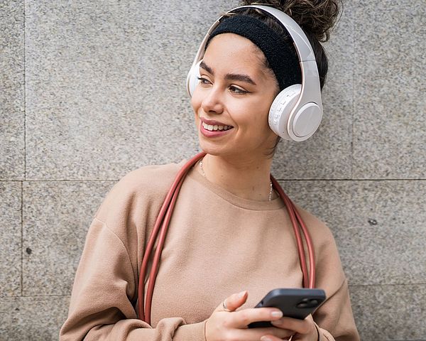 Eine junge Frau mit schwarzen, lockigen Haaren lacht und blickt zur Seite. Sie trägt weiße Over-Ear-Kopfhörer und hält ein Smartphone in beiden Händen.