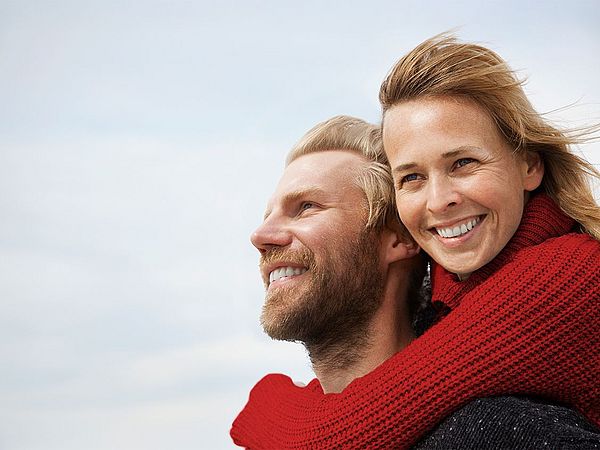 Junges Paar, bei dem die Frau den Mann umarmt, beide blicken lachend in die Ferne.