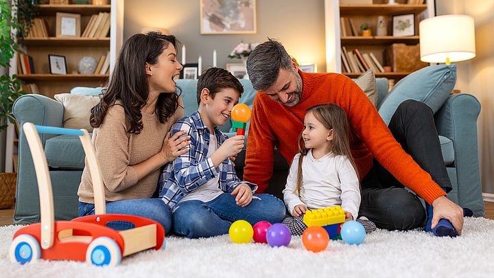 Mutter, Vater und zwei Kinder sitzen zusammen am Boden umringt von Spielsachen.