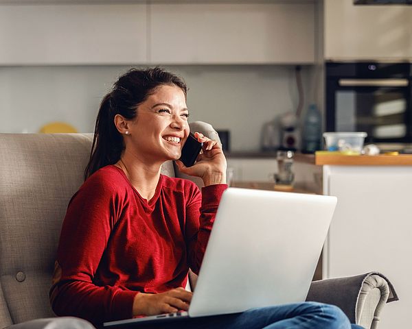 Eine glückliche Frau sitzt mit ihrem Laptop auf einem Sofa im Wohnzimmer und telefoniert